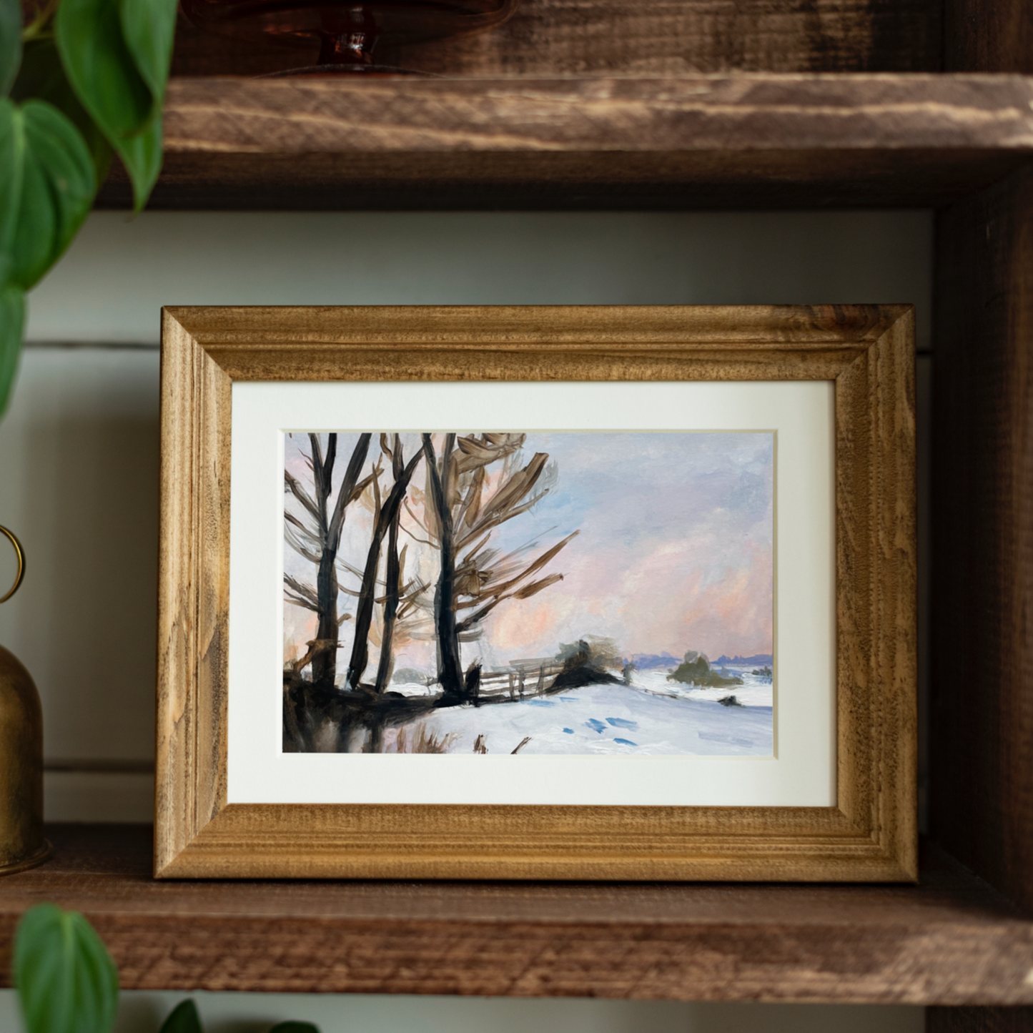 Framed winter landscape painting on a wooden shelf with plants.