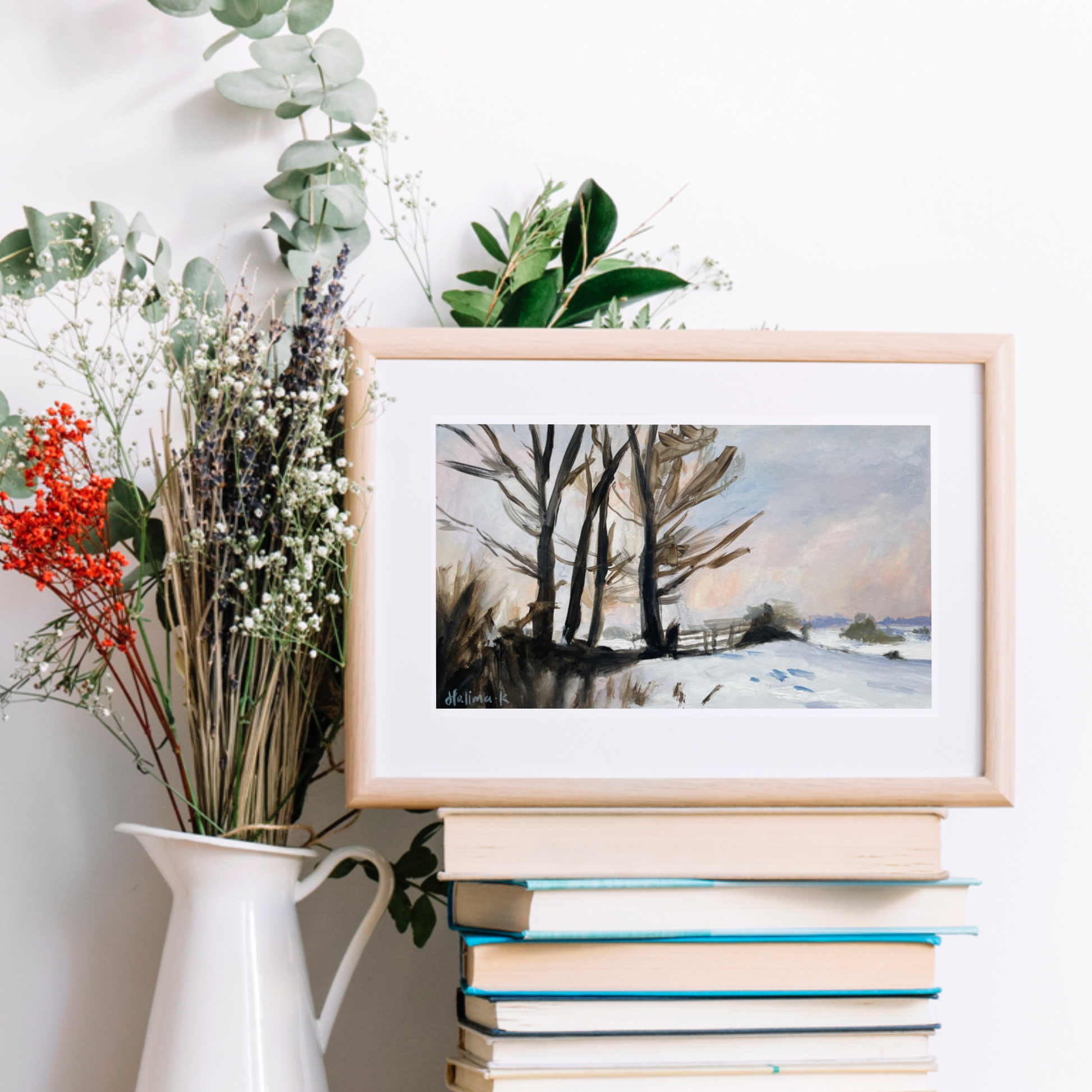 Framed winter landscape print on a stack of books with a vase of flowers.