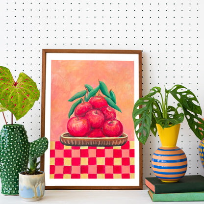 Framed artwork of apples on a checkered background with decorative plants and books on a white surface.