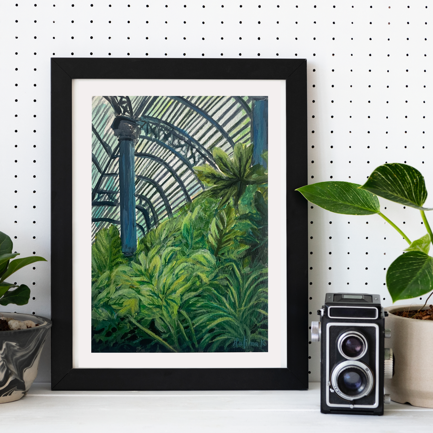Framed artwork of a lush greenhouse garden scene on a white wall with a vintage camera and planters.