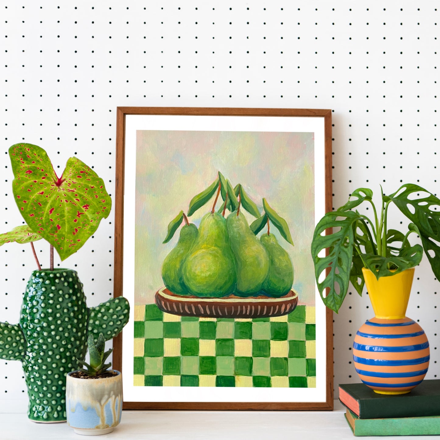 Framed artwork of pears on a checkered tablecloth with decorative plants and books in the foreground.